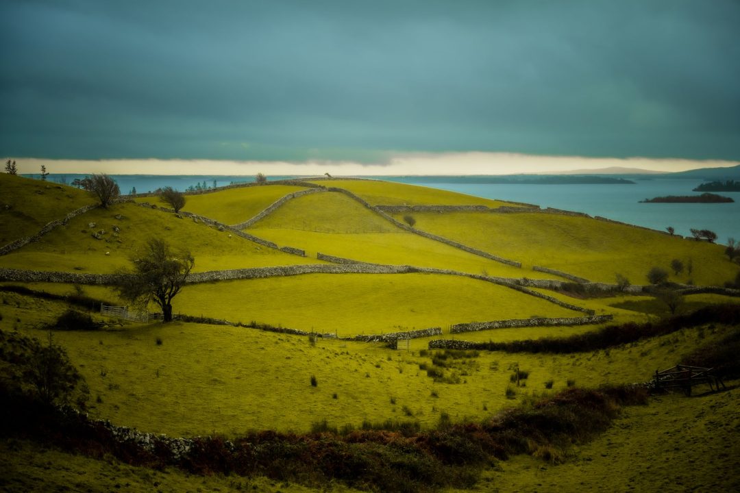 Peaceful landscape with rolling hills, stone fences, and a tranquil lake under a cloudy sky.