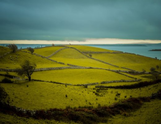 Peaceful landscape with rolling hills, stone fences, and a tranquil lake under a cloudy sky.